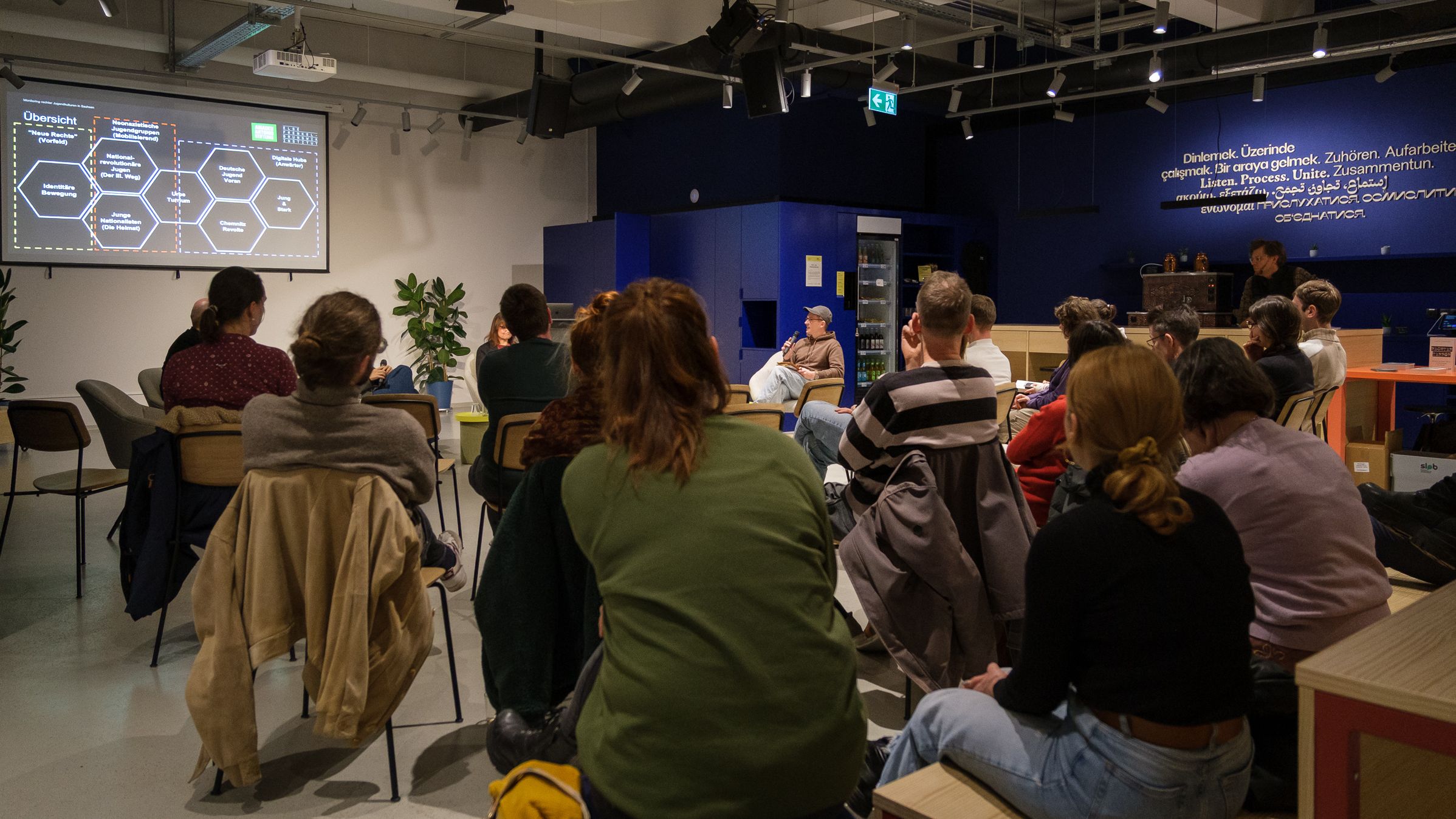 Mehrere Personen sitzen in einem Raum und schauen auf eine Präsentation mit Text und Diagrammen an der Wand.