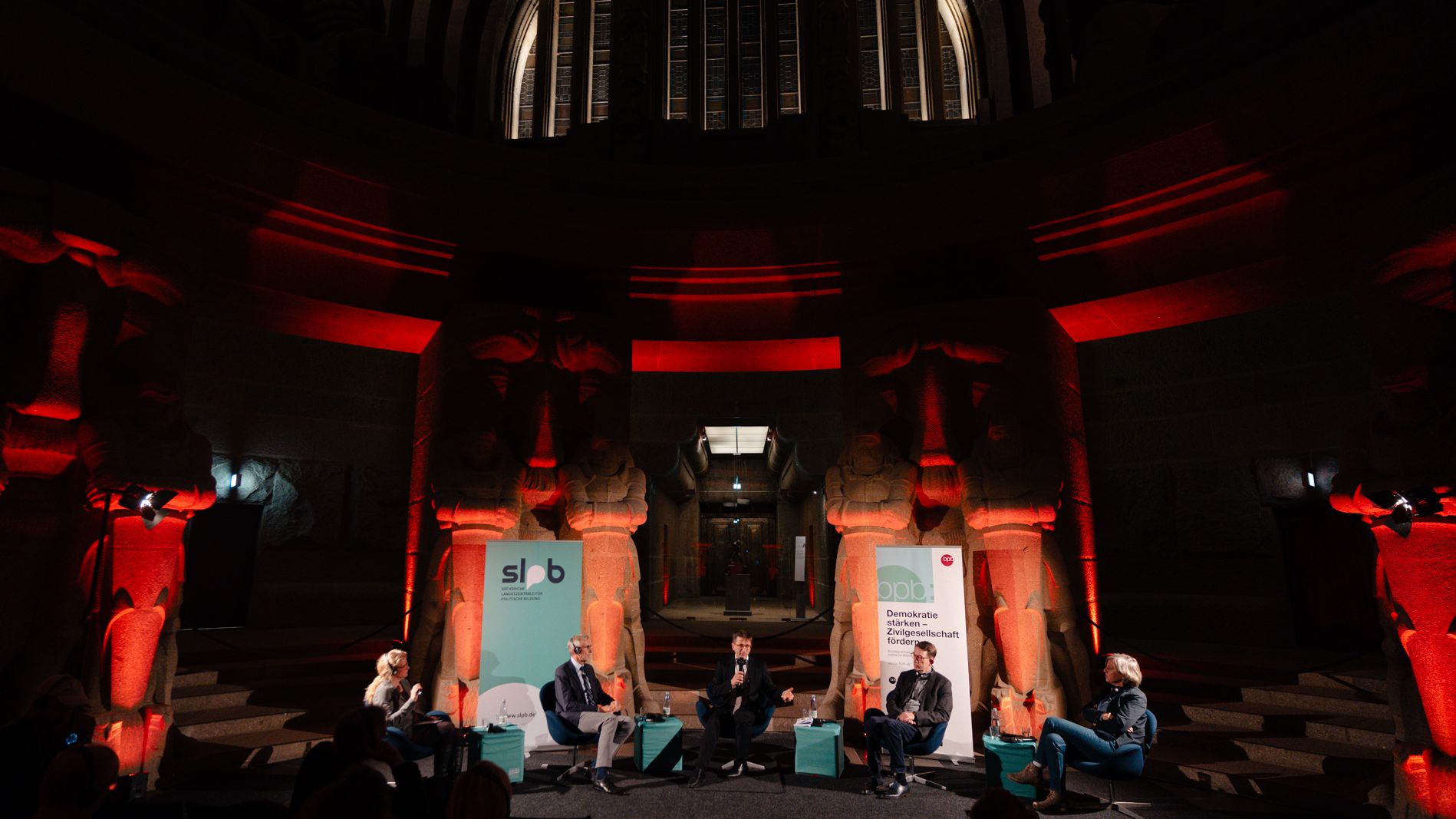 Fünf Personen sitzen auf einer Bühne im Leipziger Völkerschlachtdenkmal, umgeben von großen steinernen Skulpturen, rot beleuchtet, mit zwei Bannern im Hintergrund.