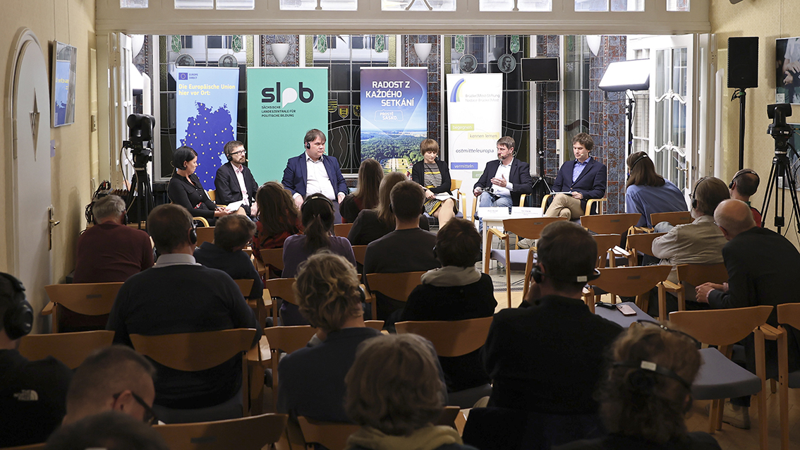 Podiumsdiskussion mit fünf Personen auf Stühlen vor Publikum in einem Raum mit hohen Fenstern und Informationsbannern im Hintergrund.