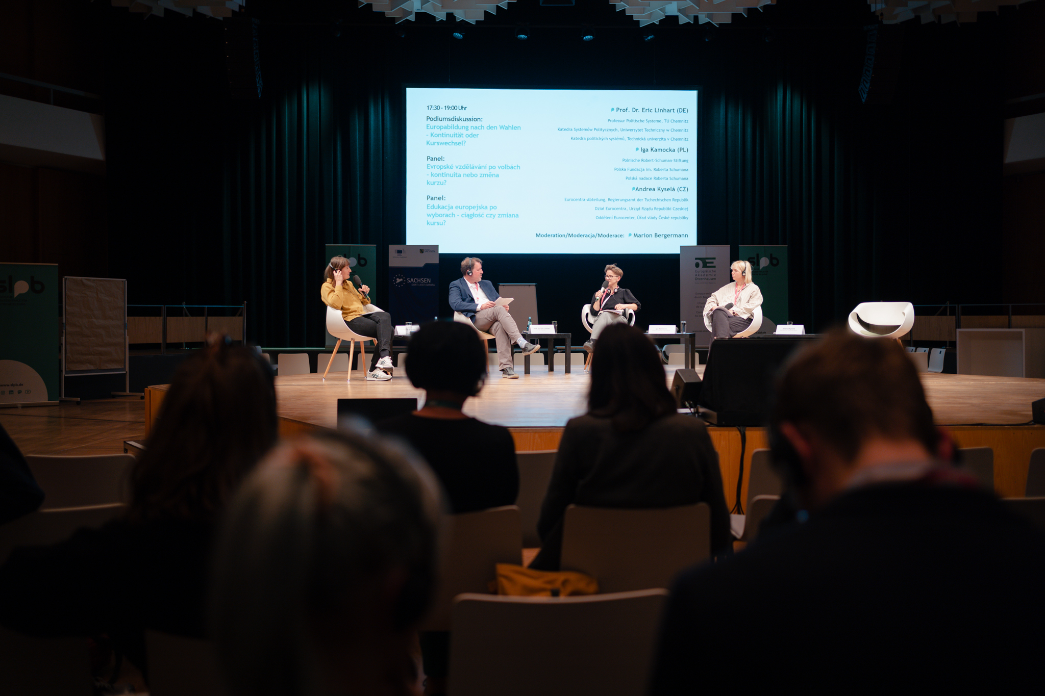 Podium mit vier Personen, drei Frauen und ein Mann, sitzen auf Stühlen auf einer Bühne vor einer großen Leinwand mit Text, Publikum im Vordergrund sichtbar.
