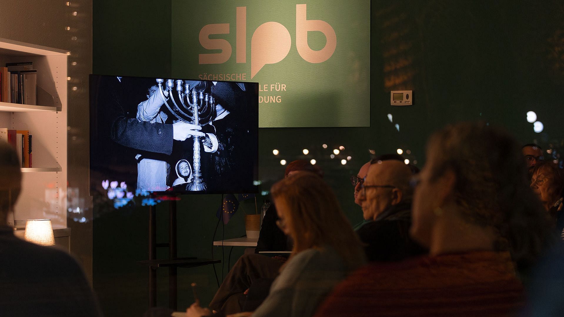 Mehrere Personen sitzen in einem dunklen Raum vor einem Bildschirm, der ein schwarz-weißes Bild zeigt von Personen die einen jüdischen Kerzenleuchter tragen, im Hintergrund ein Schild mit der Aufschrift 'slpb'.