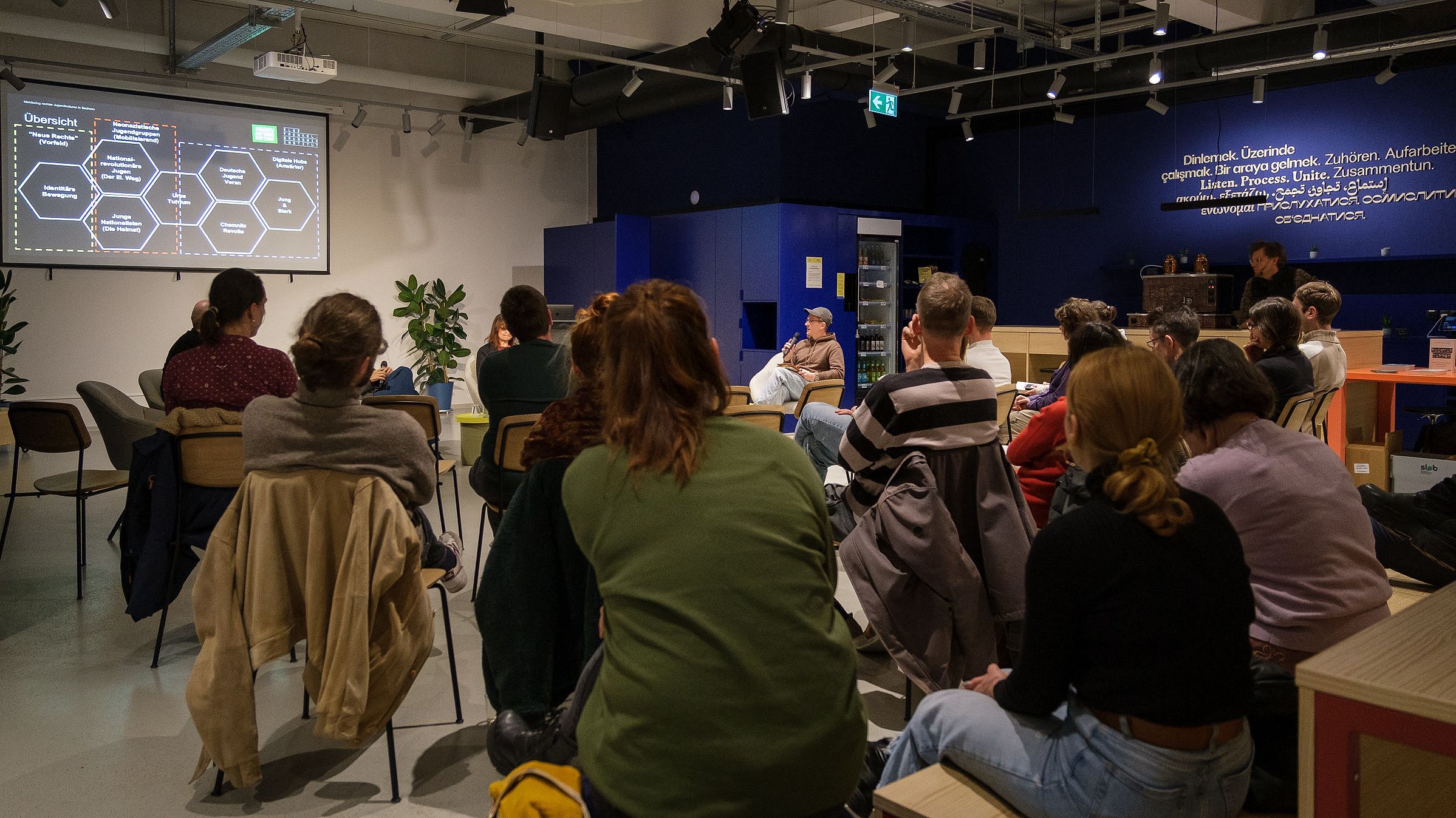 Mehrere Personen sitzen in einem Raum und schauen auf eine Präsentation mit Text und Diagrammen an der Wand.