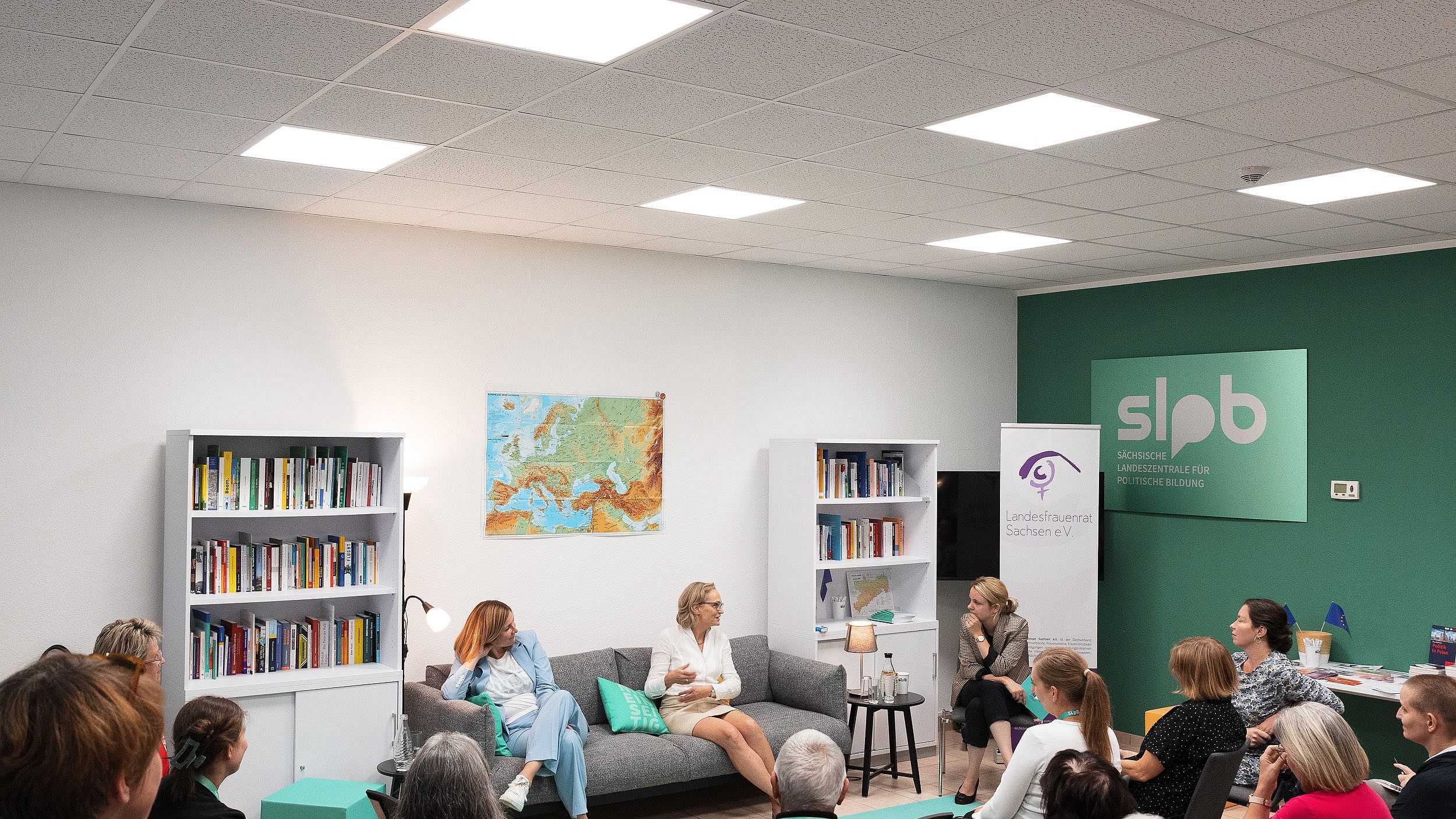 Zwei Frauen sitzen auf einem grauen Sofa in einem Raum mit Bücherregalen. Eine Frau hält ein Mikrofon, während die andere spricht. Im Hintergrund steht ein Schild mit 'slpb'.