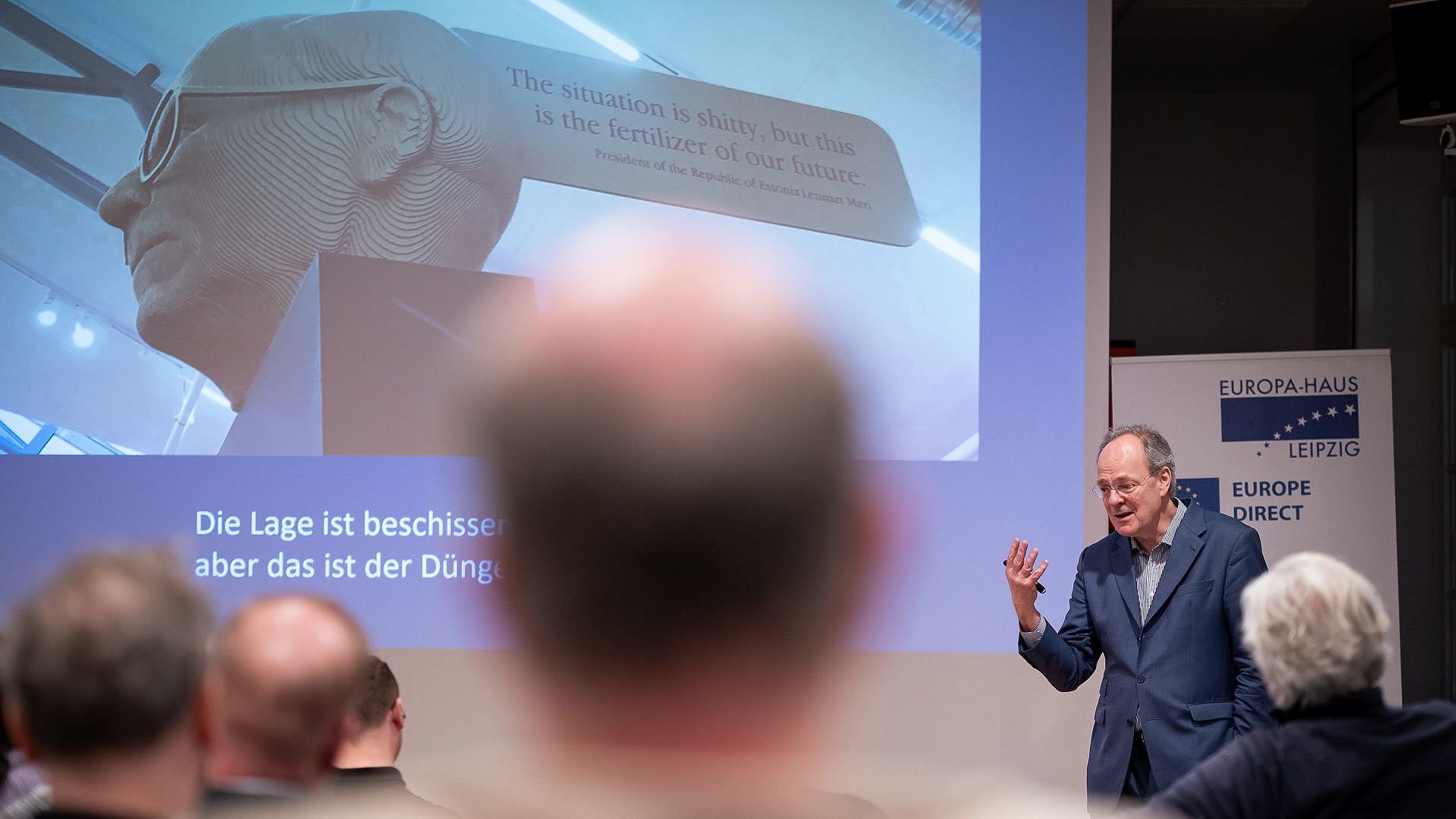 Ein Mann im Anzug hält eine Präsentation vor einem Publikum mit einer Projektion, auf der englischer und deutscher Text zu sehen ist.