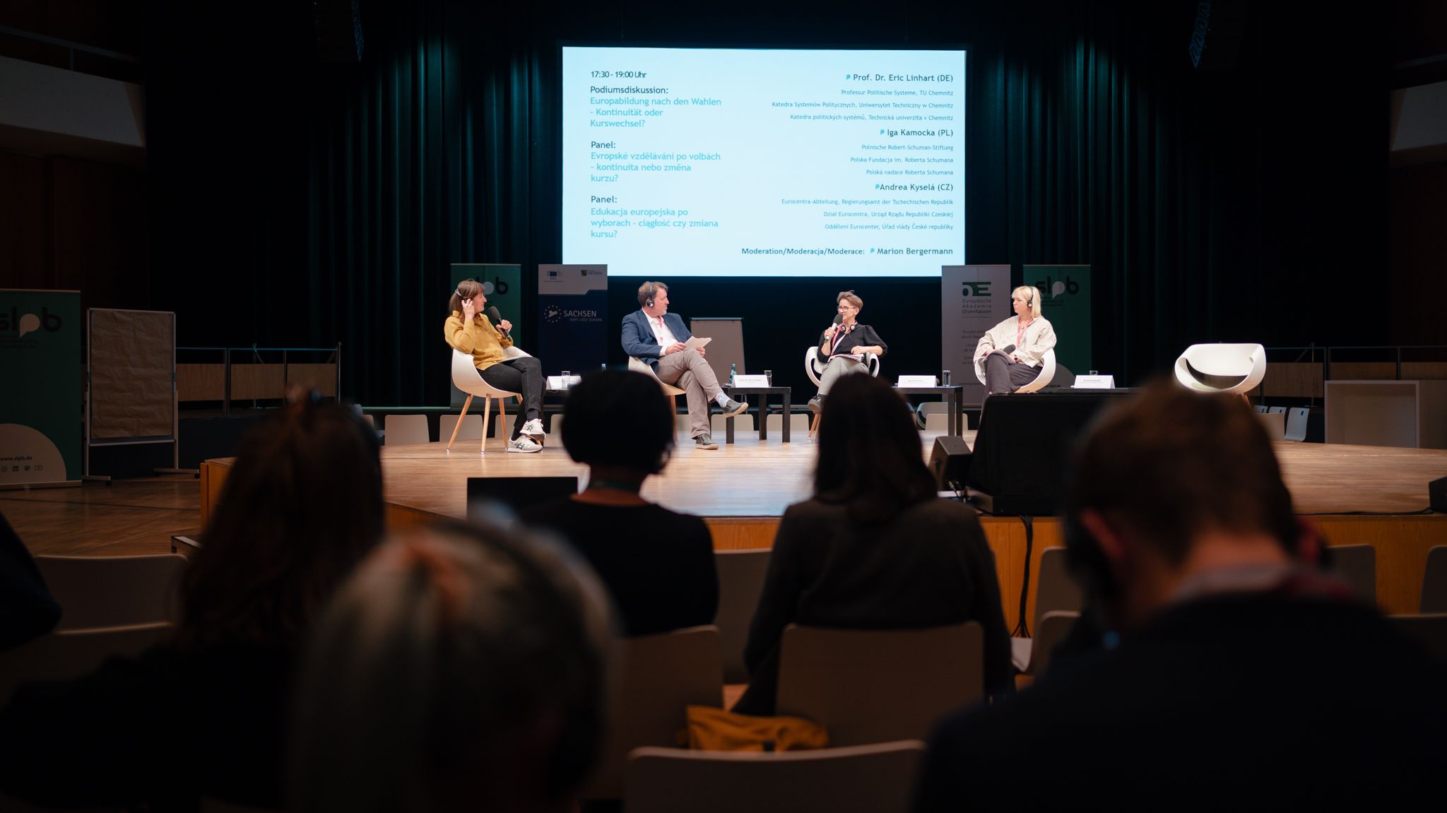 Podium mit vier Personen, drei Frauen und ein Mann, sitzen auf Stühlen auf einer Bühne vor einer großen Leinwand mit Text, Publikum im Vordergrund sichtbar.