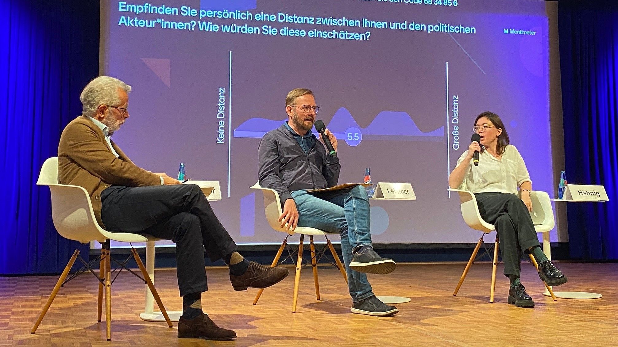 Theodor-Litt-Symposium Das Bild zeigt eine Podiumsdiskussion mit drei Personen, die auf Stühlen auf einer Bühne sitzen. Im Hintergrund ist eine große Leinwand mit einer Grafik und Text, der auf Deutsch lautet: "Empfinden Sie persönlich eine Distanz zwischen Ihnen und den politischen Akteur*innen? Wie würden Sie diese einschätzen?" Darunter befinden sich Skalenangaben mit den Beschriftungen "Keine Distanz" und "Große Distanz", sowie eine Zahl "5.5", die vermutlich eine Durchschnittsbewertung darstellt. Namenstafeln sind vor den Teilnehmenden platziert, allerdings ist nur der Name "Hämig" vollständig sichtbar. Bei Klick vergrößert sich das Bild.