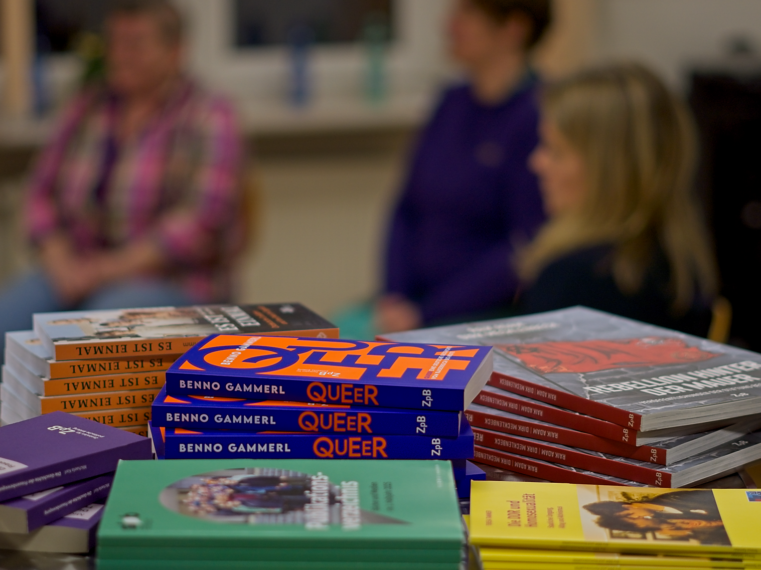 Im Hintergrund sind die drei Podiumsgäste unscharf zu erkennen. von links: Elke Prinz, Barbara Wallbraun und Teresa Tammer. Im Vordergrund ist scharf ein Büchertisch zu erkennen.