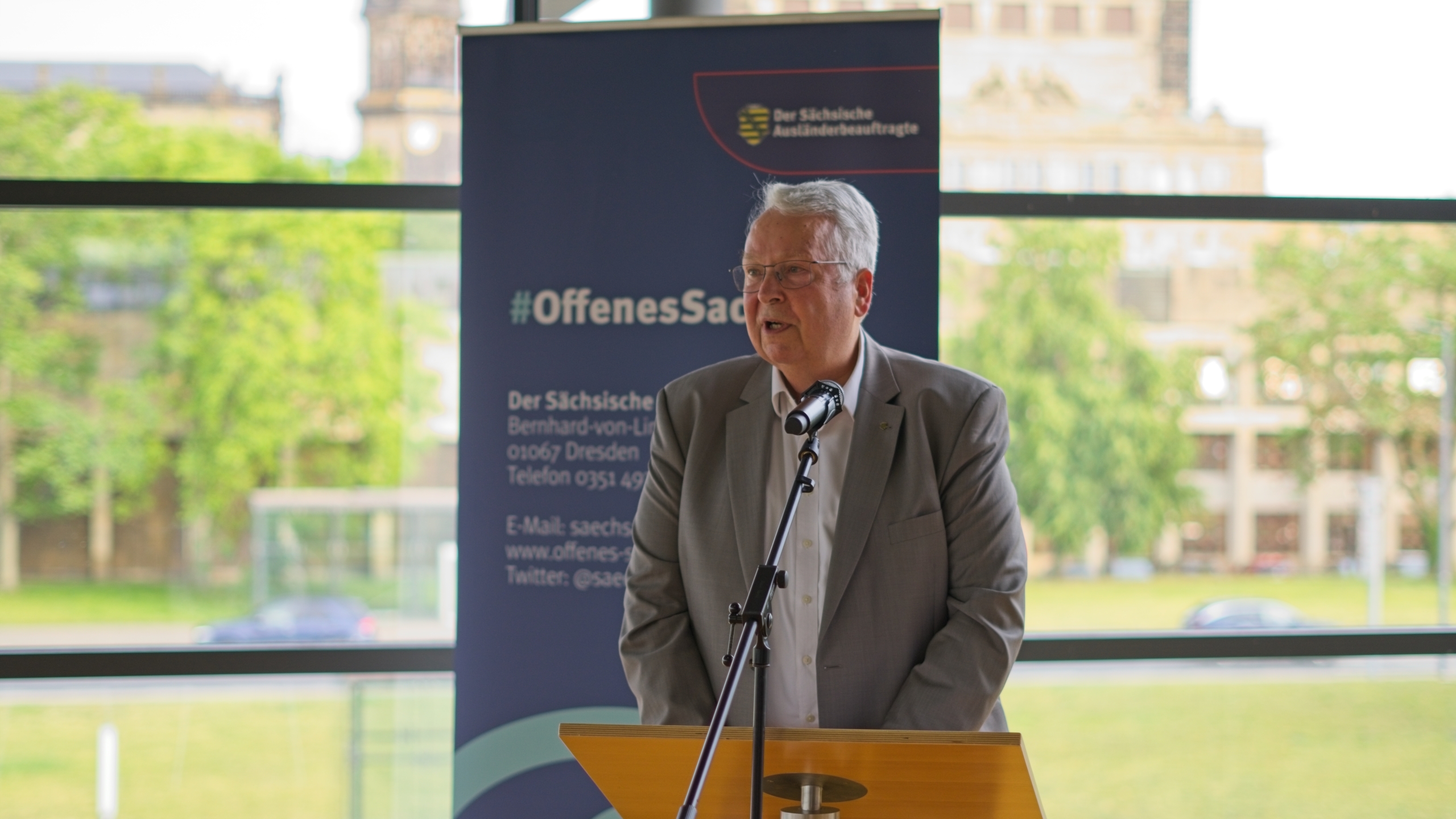 Geert Mackenroth spricht bei der Feier zum 20-jährigen Bestehen der Sächsischen Härtefallkommission vor einem Mikrofon mit Banner im Hintergrund