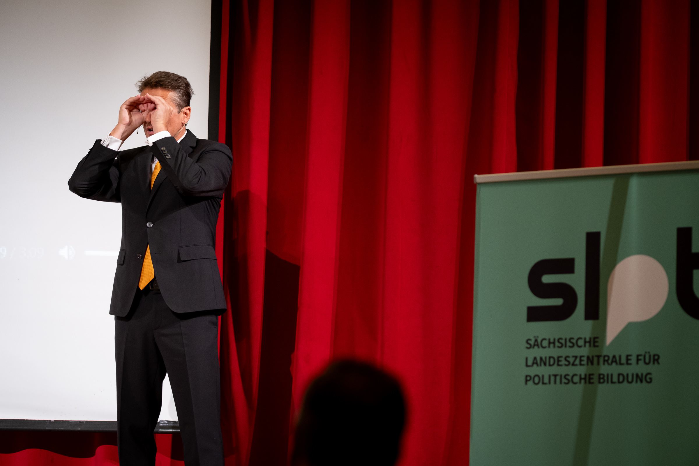 Mann im schwarzen Anzug mit orangefarbener Krawatte steht vor rotem Vorhang und weißer Leinwand, rechts ein Banner mit Schriftzug 'slpb Sächsische Landeszentrale für politische Bildung'.