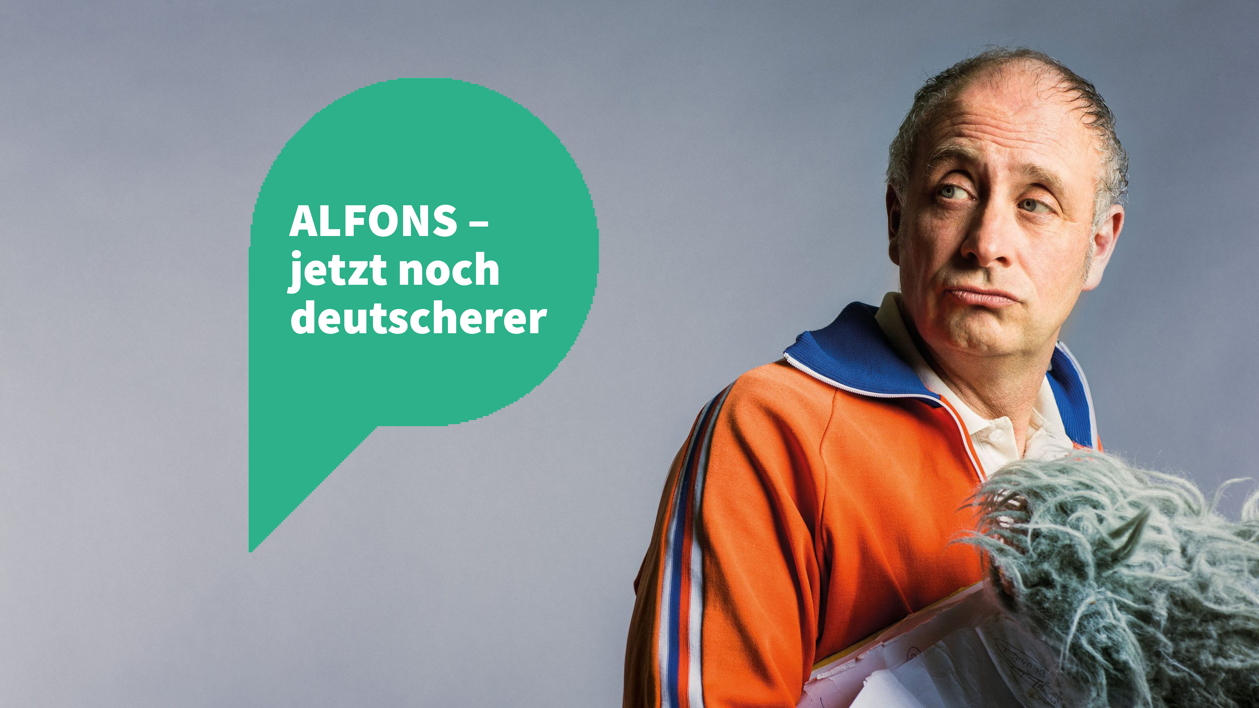 Rechts im Bild steht Alfons in einer orangenen Traningsjacke und schaut sich über die rechte Schulter. links um Bild ist eine grüne Sprechblase mit dem Text "Alfons - jetzt noch deutscher"